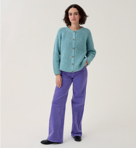 Woman wearing a Sirdar 10780 teal knitted cardigan and purple pants on a white background