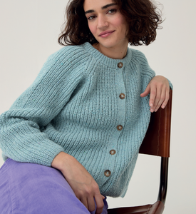 Woman wearing a light blue knitted cardigan with brown buttons, sitting on a chair against a plain background - Sirdar 10780 Loveful 100% Recycled Tweed Double Knit Ribbed Cardigan Knitting Pattern