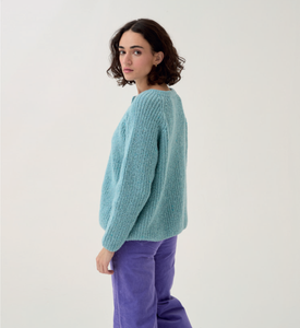 Woman wearing a light blue sweater and purple pants on a white background - Sirdar 10780 Loveful 100% Recycled Tweed Double Knit Ribbed Cardigan Knitting Pattern