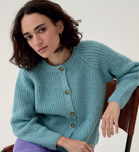 Woman wearing a Sirdar 10780 Loveful 100% Recycled Tweed Double Knit ribbed cardigan sitting on a chair against a white background