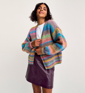 Woman wearing a colourful Sirdar 10801 Jewelspun Chunky knitted cardigan over a white shirt and purple skirt on a light gray background